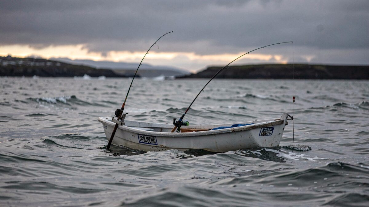 A small open boat drifting on a grey north coast swell at first light, a mooching rod bent in a holder, a cut-plug herring rig visible on the surface, low cloud over a dark shoreline.